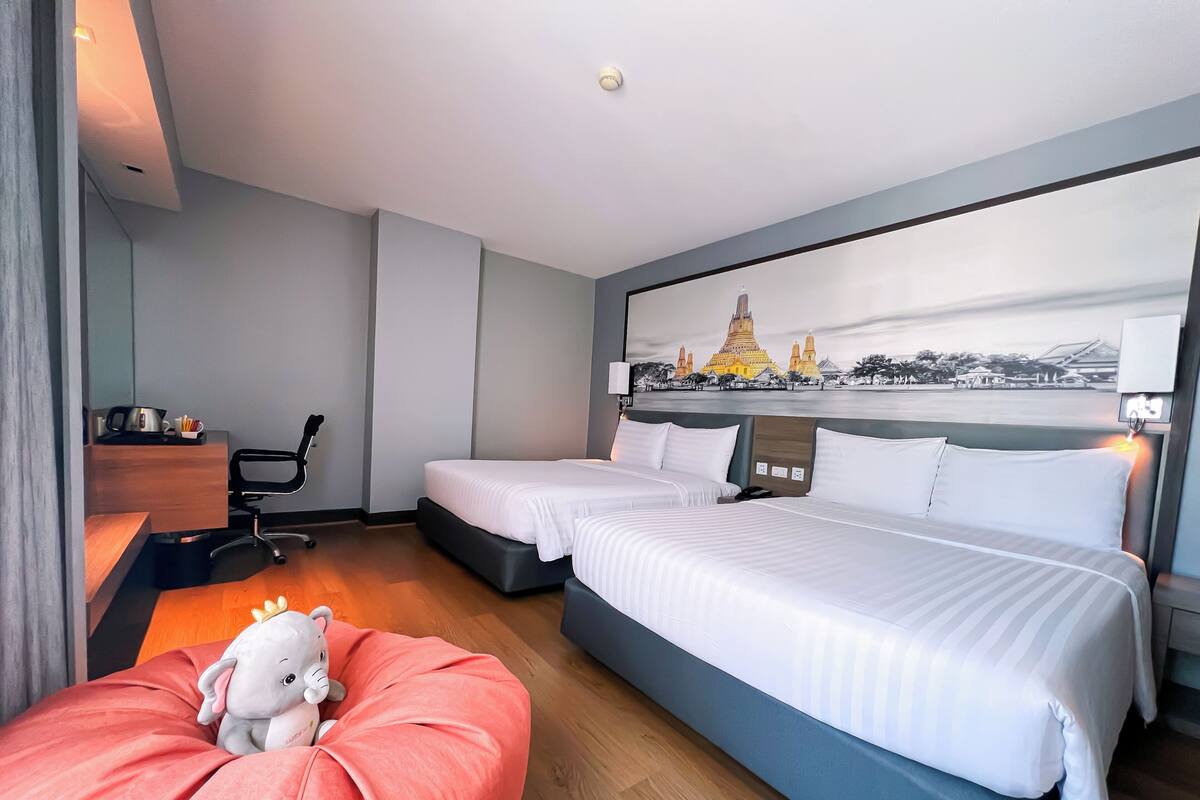 Habitación de hotel con dos camas, un escritorio, una silla, un sillón de mimbre rojo y una lámpara fijada a la pared.