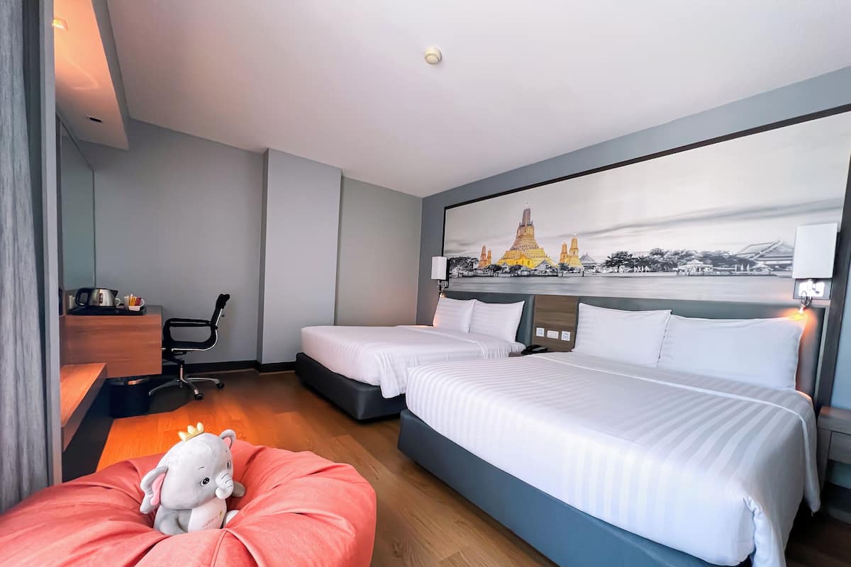 A hotel room with two beds, a desk, a chair, a red bean bag, and a wall-mounted lamp.