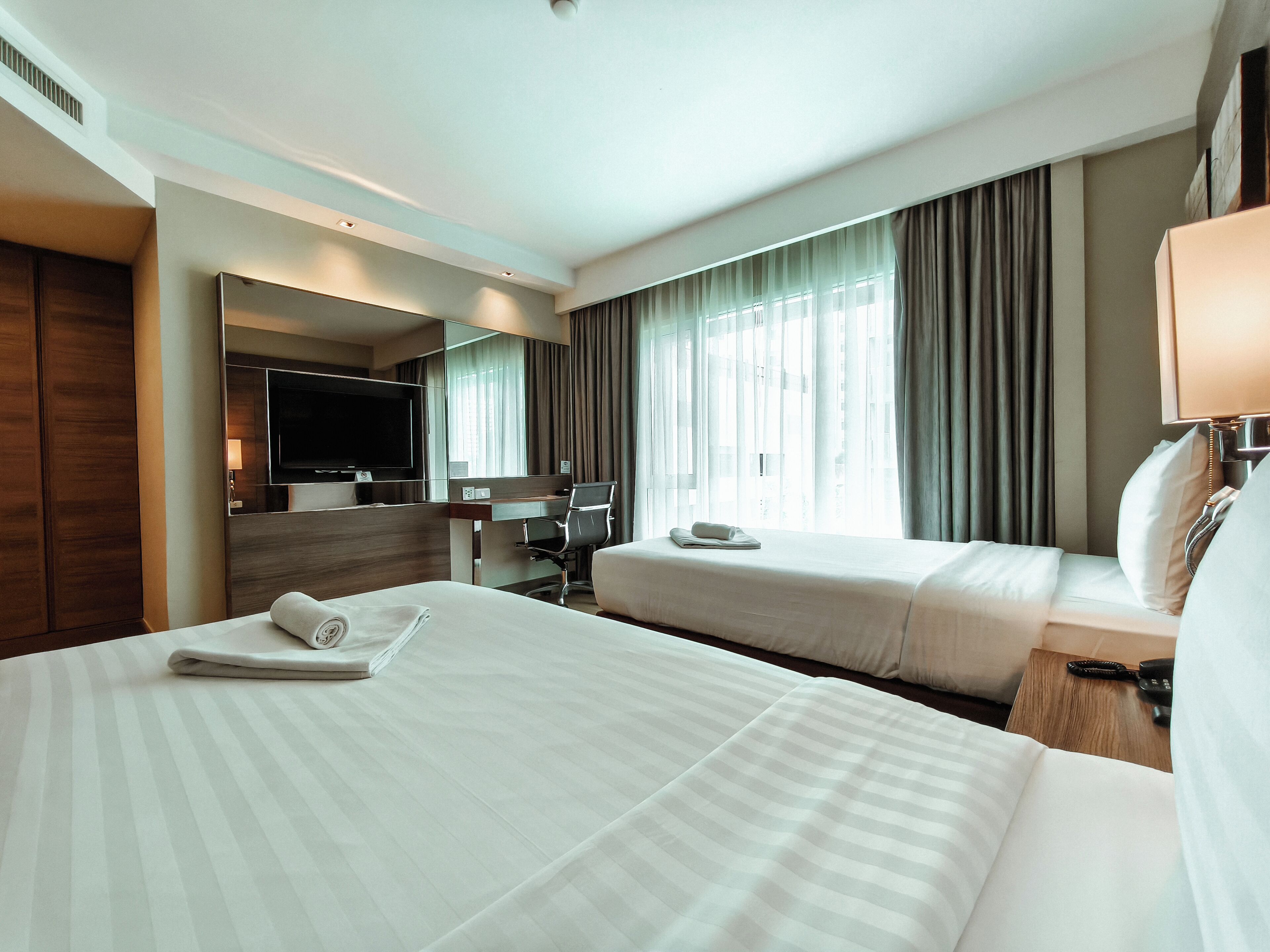 Ein Hotelzimmer mit zwei Betten, einem Schreibtisch, einem Sessel, einem Fernseher und einem großen Fenster mit Vorhängen.
