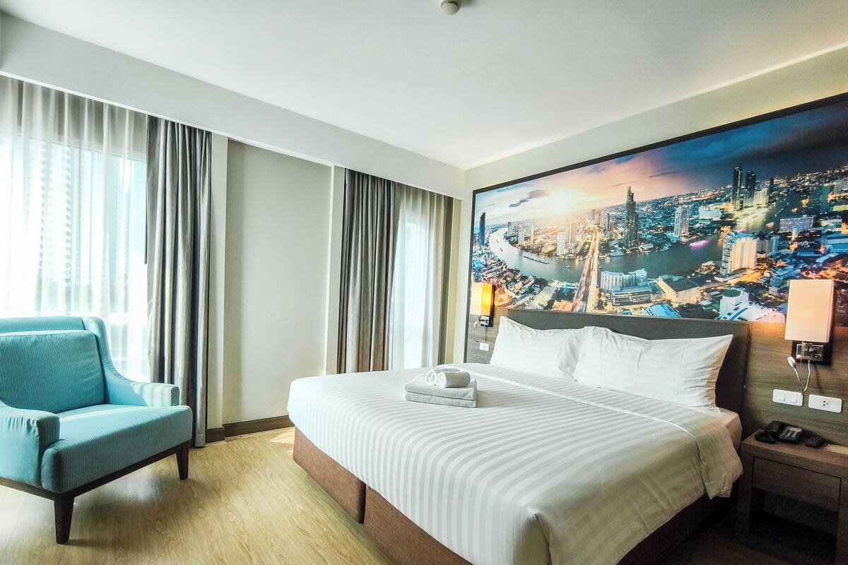 Habitación de hotel con una cama grande, un sillón azul, un mural con vista urbana en la pared y una mesita de noche con teléfono y lámpara.