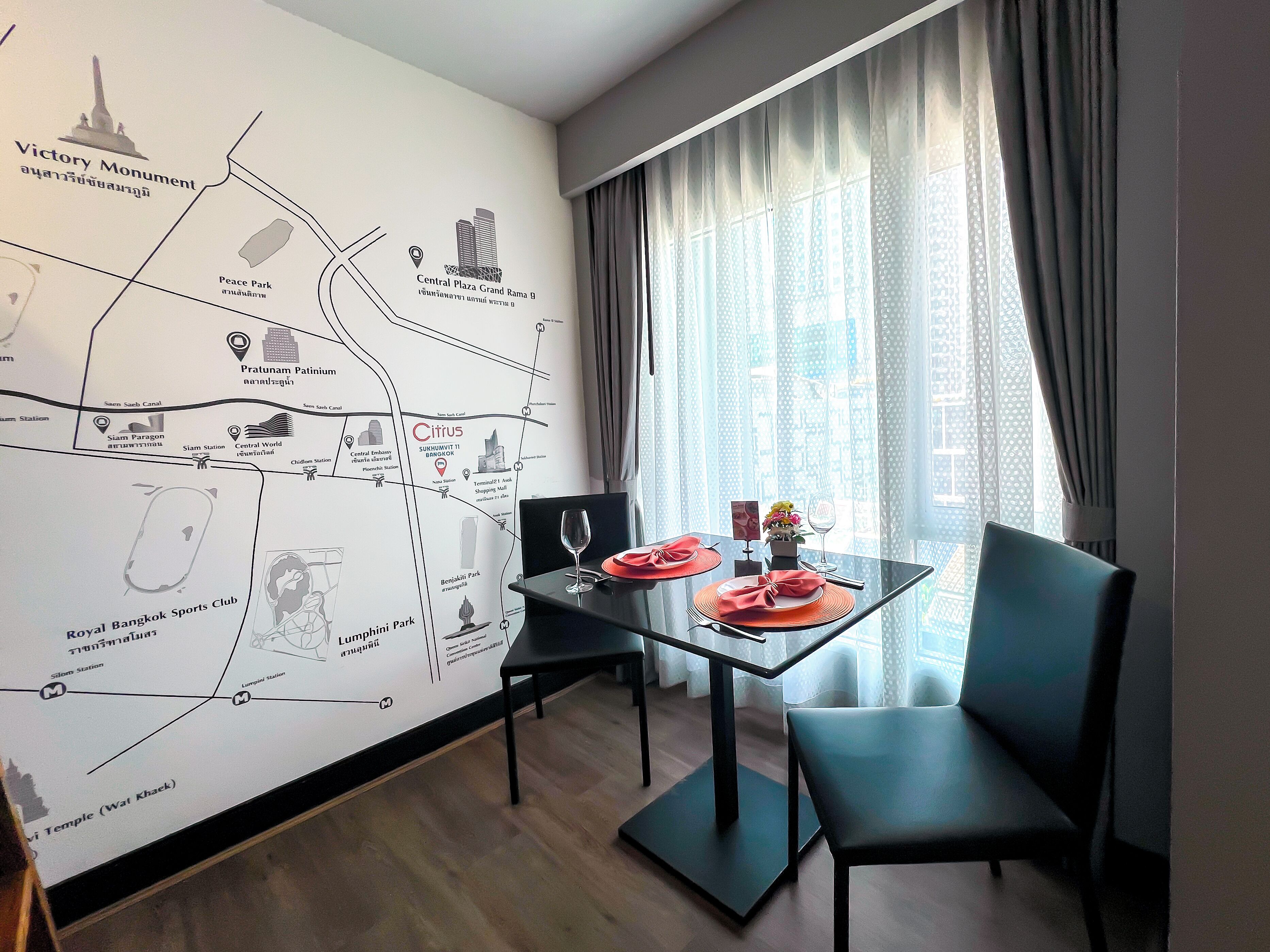 Een hotelkamer met een wand vol kaarten, een eettafel met stoelen, een glazen scheiding en uitzicht op de stad door een raam.