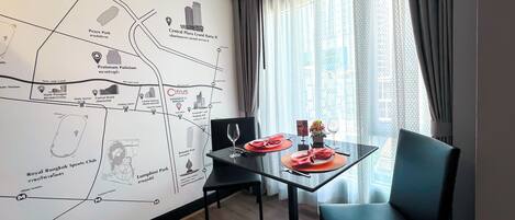 Een hotelkamer met een wand vol kaarten, een eettafel met stoelen, een glazen scheiding en uitzicht op de stad door een raam.