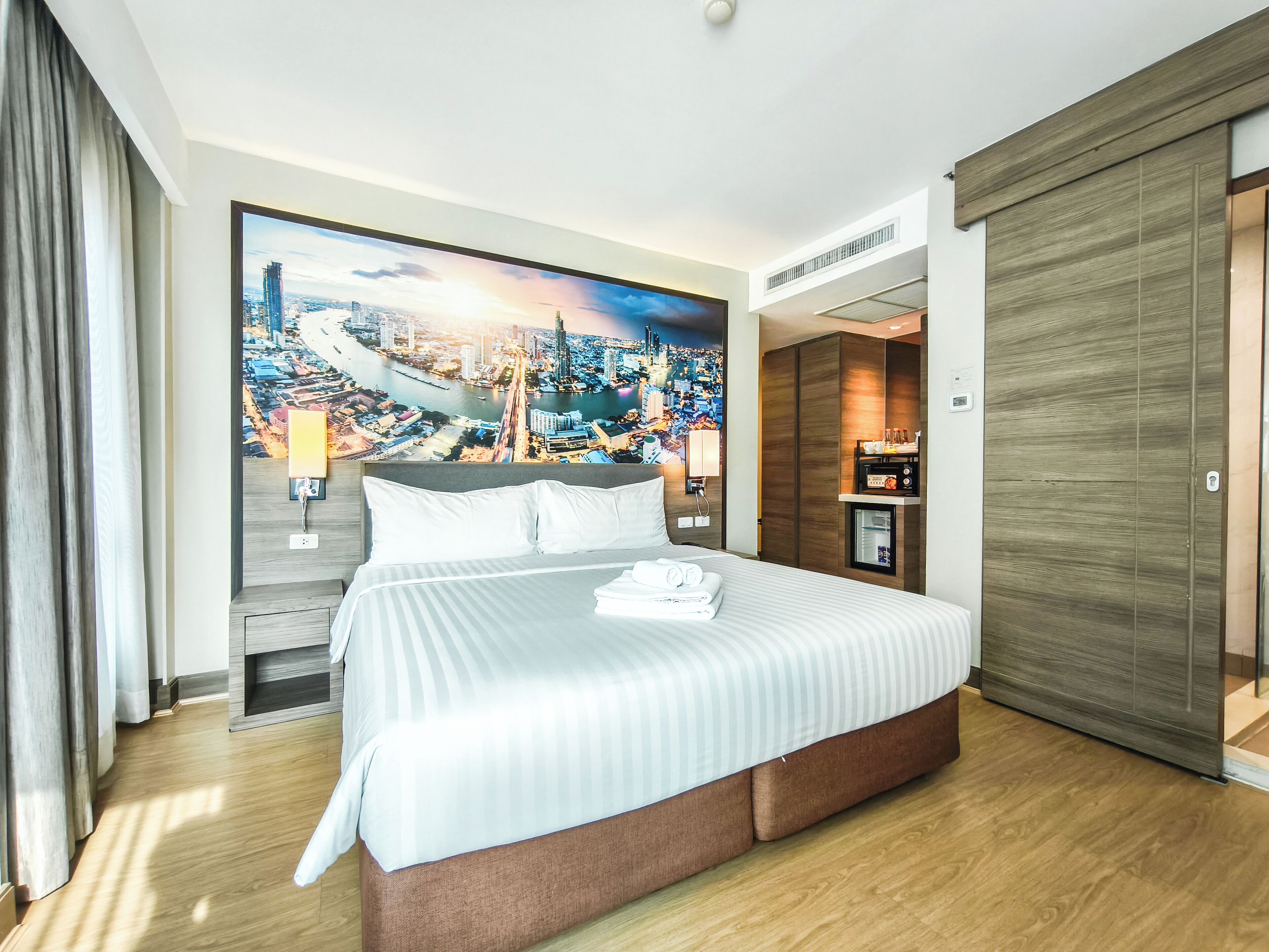 A hotel room with a large bed, a cityscape mural on the wall, a minibar, and a wooden wardrobe.
