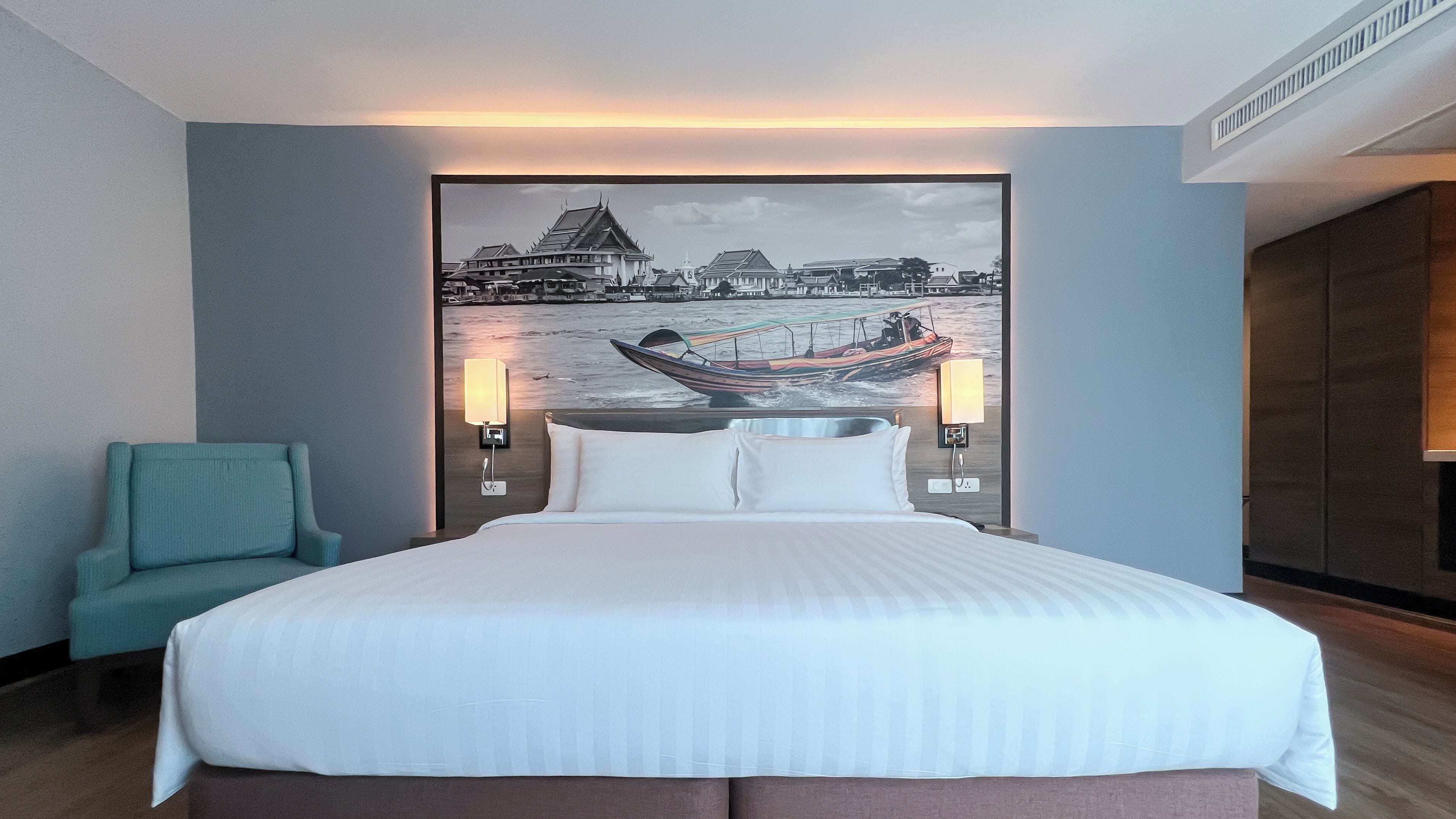 Una habitación de hotel con una cama grande, una silla, un cuadro colgado en la pared que muestra un barco y edificios, y dos lámparas de pared.