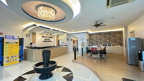 Zona de recepción de hotel con una lámpara de araña moderna, un mostrador de recepción, un área de comedor con una mesa redonda y sillas, y una cocina con refrigerador y microondas.