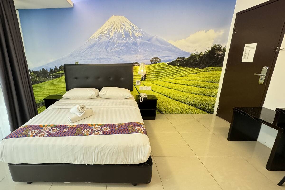 Habitació d'hotel amb llit, una taula de nit, una làmpada i un mural a la paret amb una muntanya.