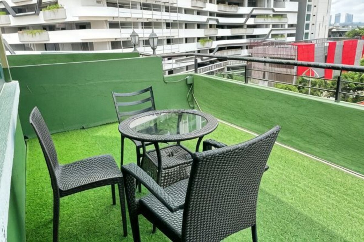 A balcony with a round table and four chairs, surrounded by green artificial grass, with a view of a multi-story building in the background.