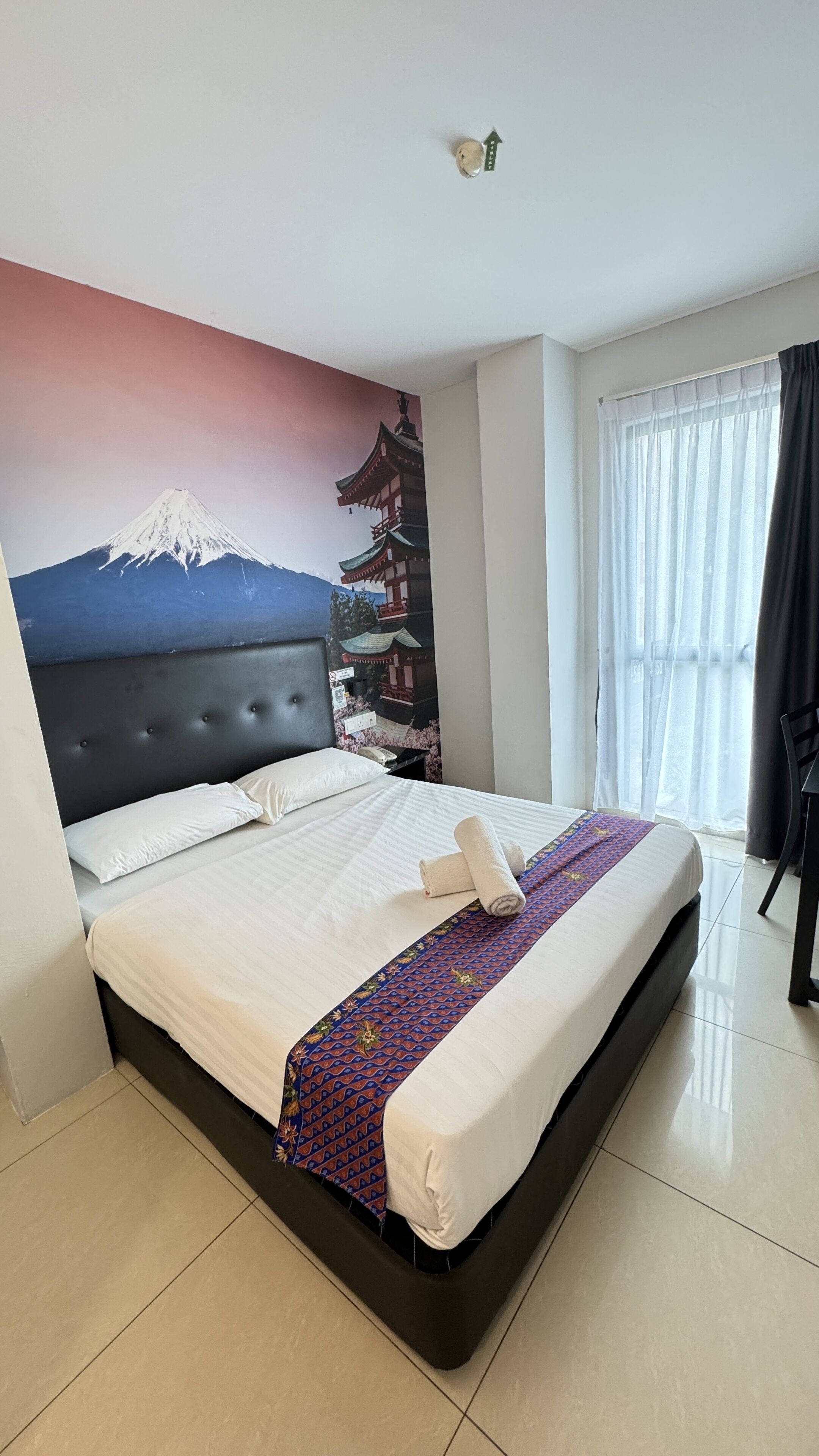 Ein Hotelzimmer mit Bett, Wandgemälde von Berg und Pagode, einem Fenster mit Vorhängen und einem Fliesenboden.