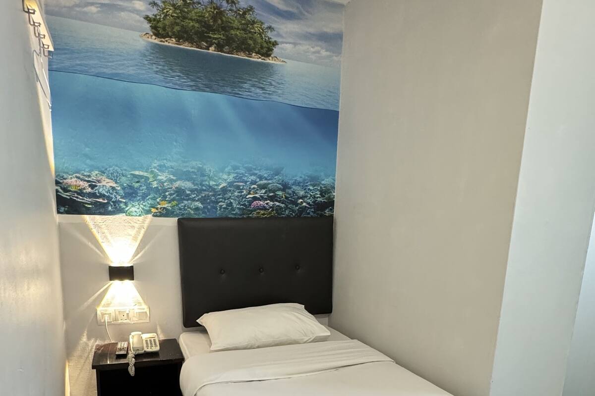 Una habitació d'hotel amb un llit, una tauleta de nit amb una làmpada i un mural a la paret que representa una illa tropical.
