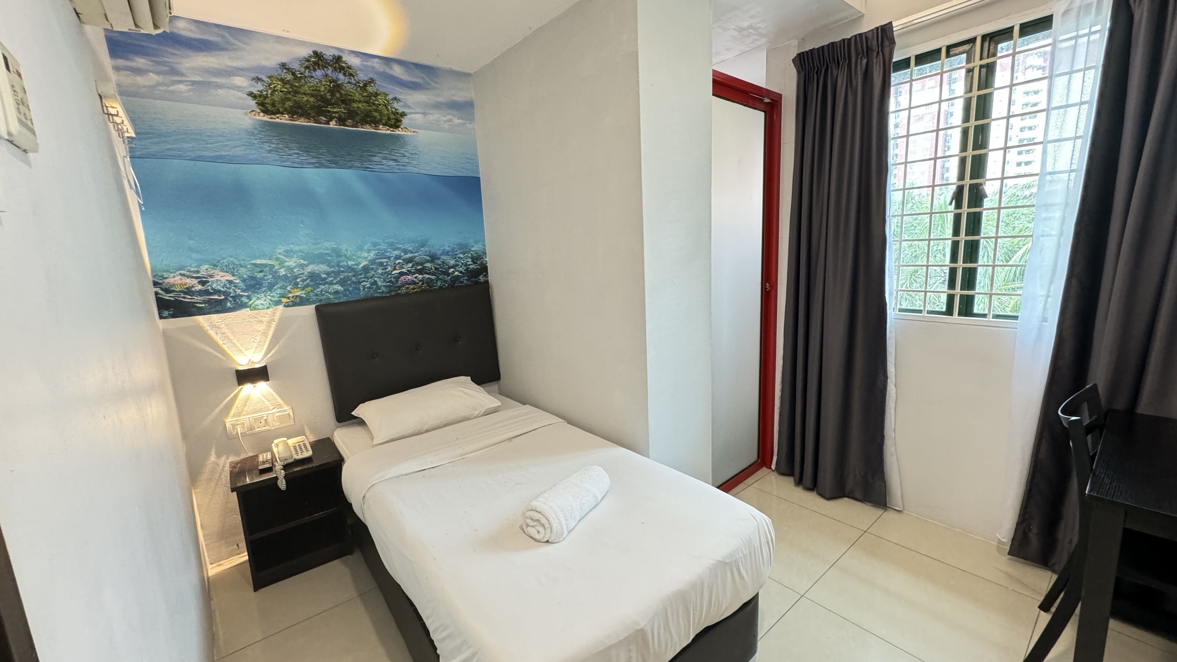 A hotel room with a bed, a nightstand, a lamp, a window with curtains, and a painting of a tropical island.