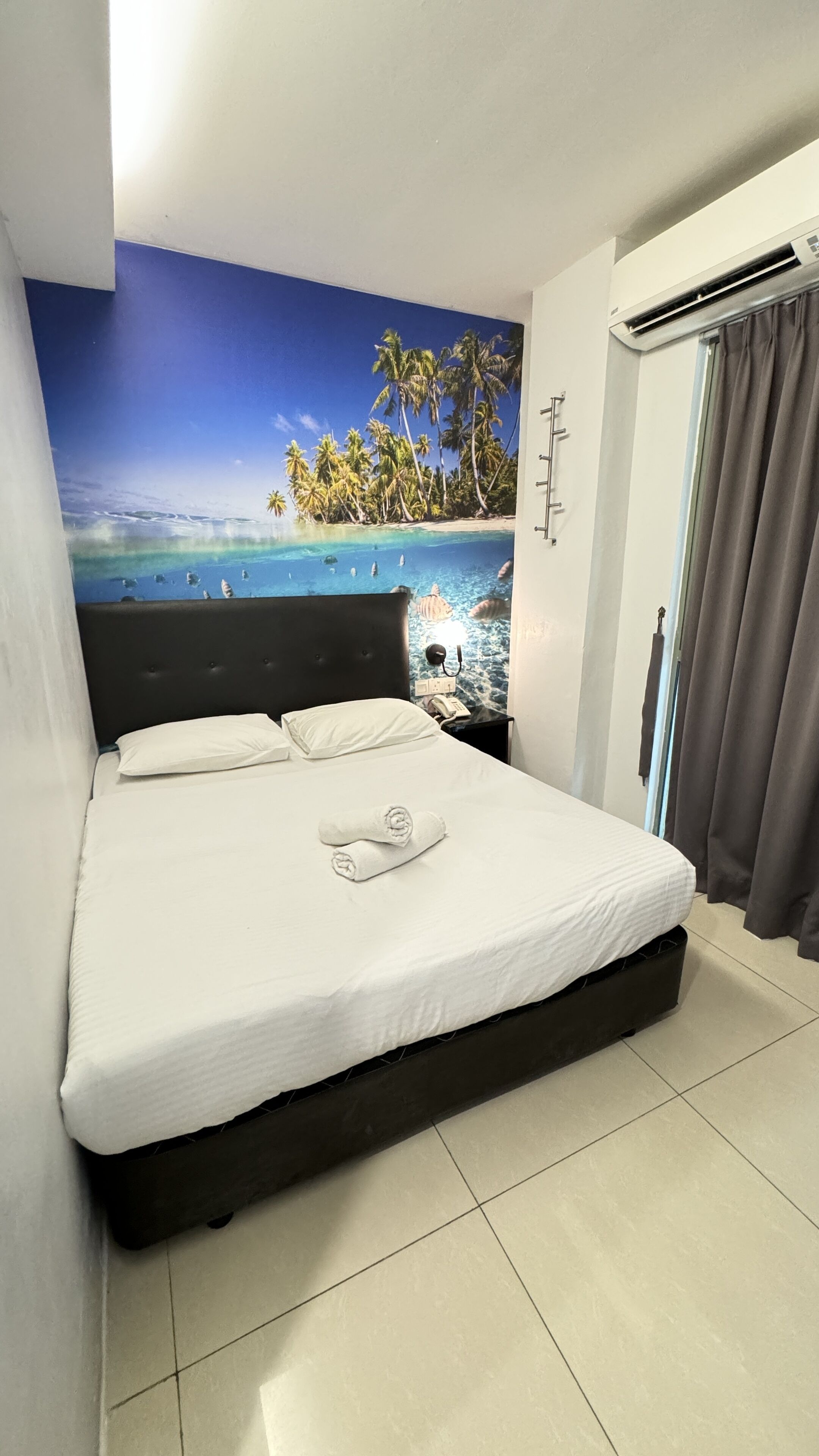 A bedroom with a bed, a nightstand, a wall mural of a beach, and an air conditioning unit.