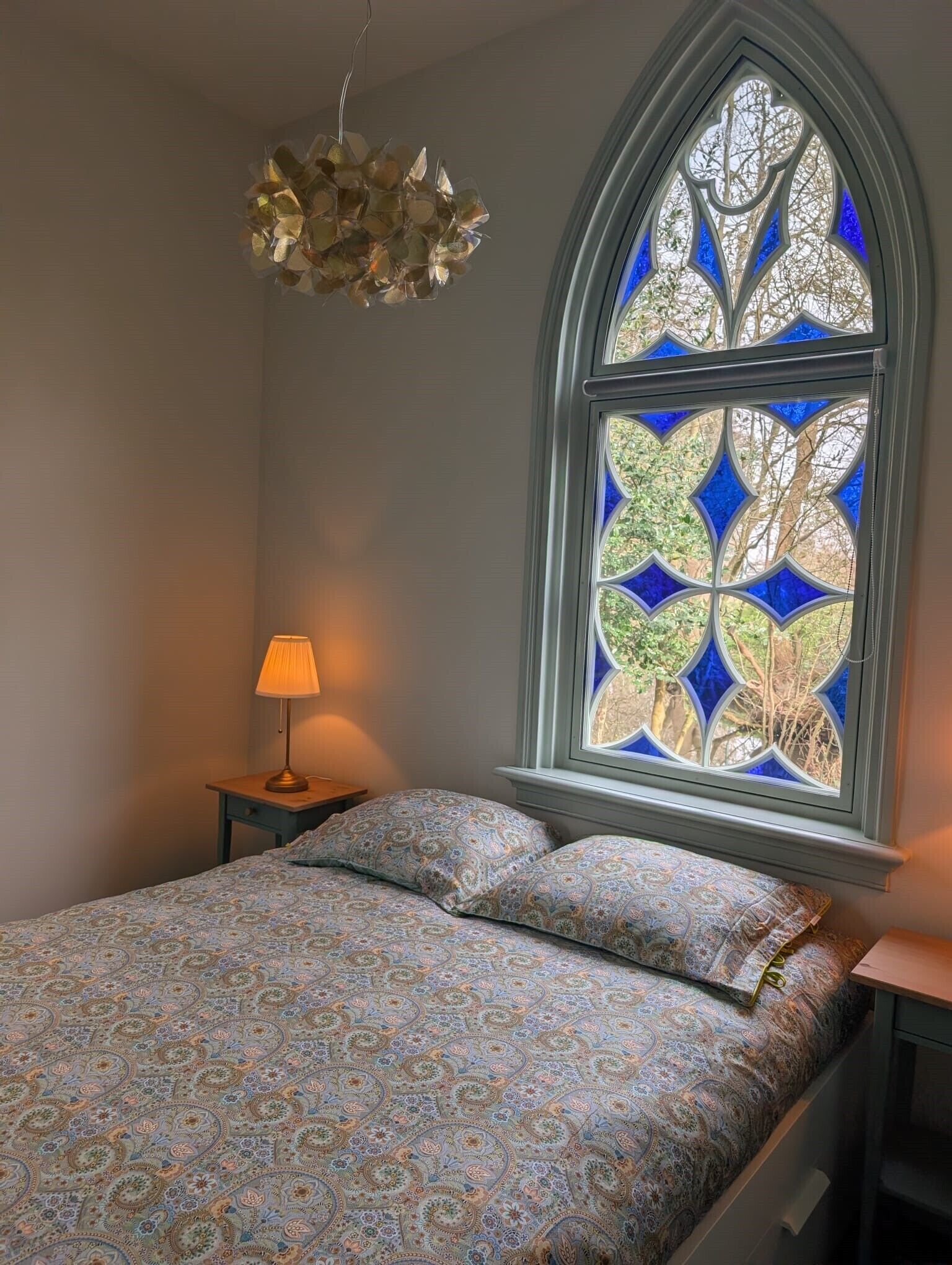 A bedroom with a bed, a nightstand, a lamp, and a stained glass window.