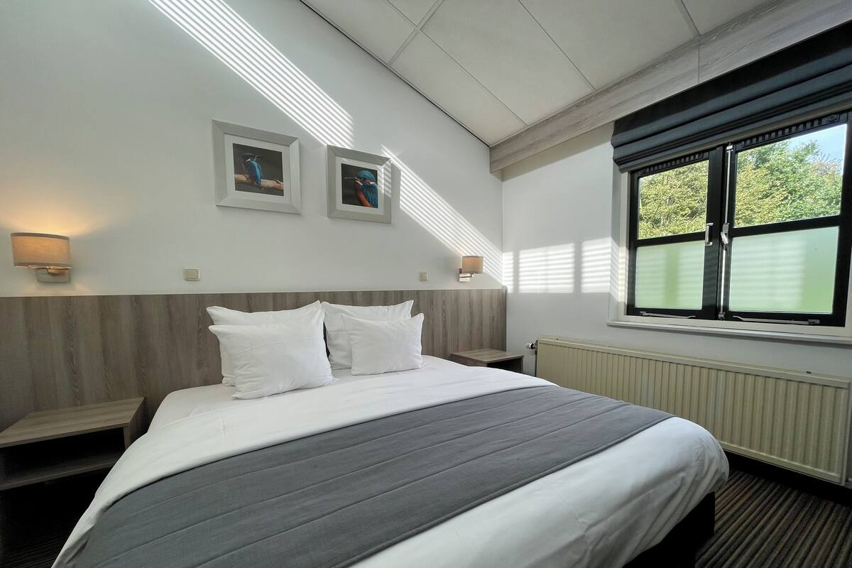 A hotel room with a large bed, two bedside tables, a window with blinds, and two wall-mounted lamps.