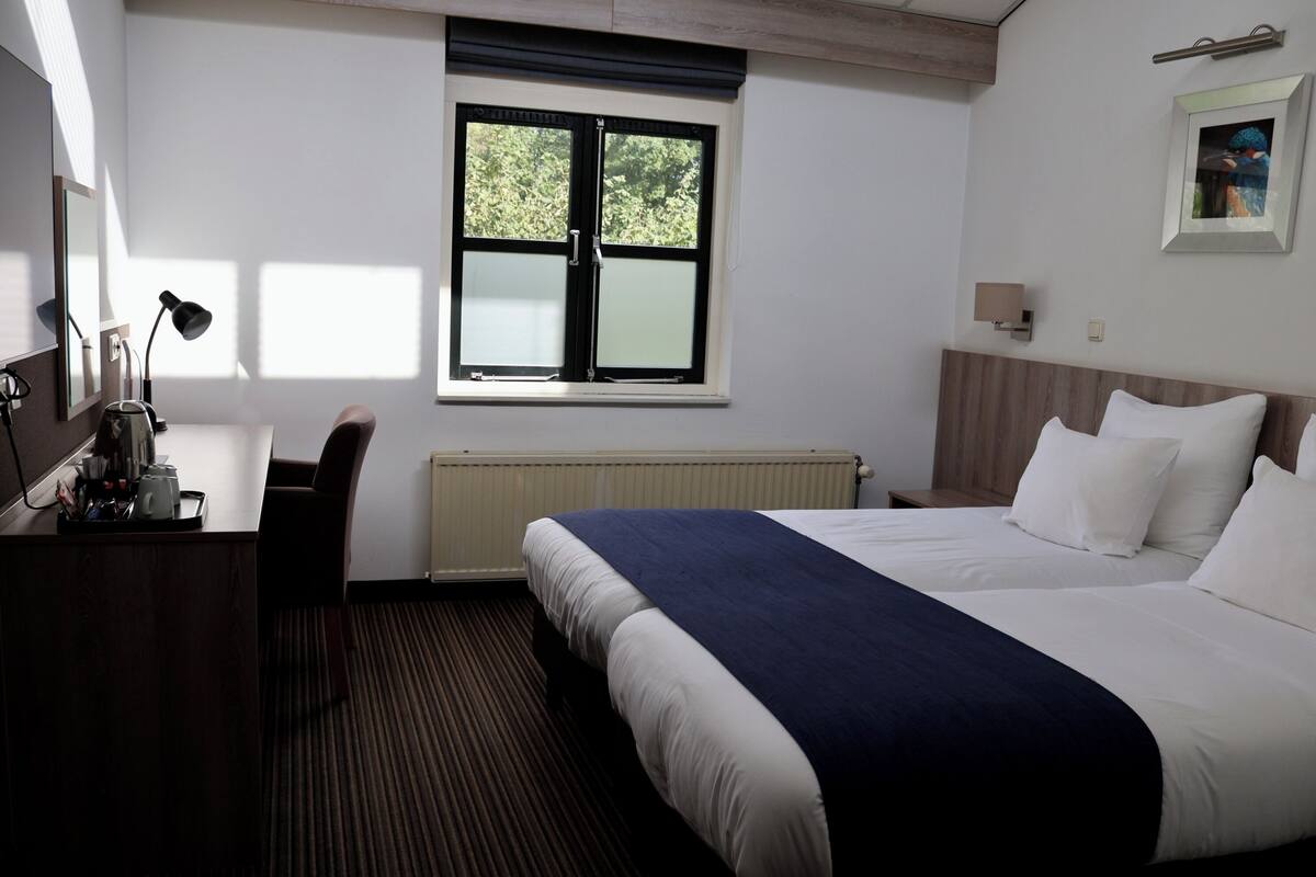 A hotel room with a bed, desk, chair, and a window with blinds.