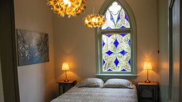 A bedroom with a bed, two bedside tables with lamps, a painting on the wall, and a stained glass window.