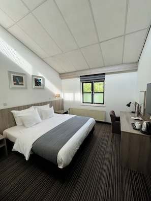 Een hotelkamer met een groot bed, een bureau met stoel, een raam met jaloezieën en twee ingelijste foto's aan de muur.