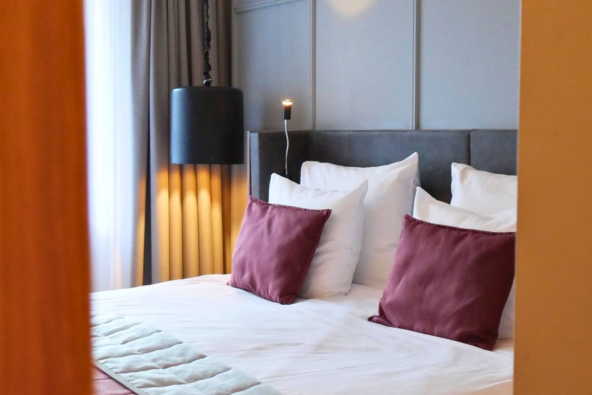 A hotel room with a large bed, a headboard with a light fixture, and a window with curtains.