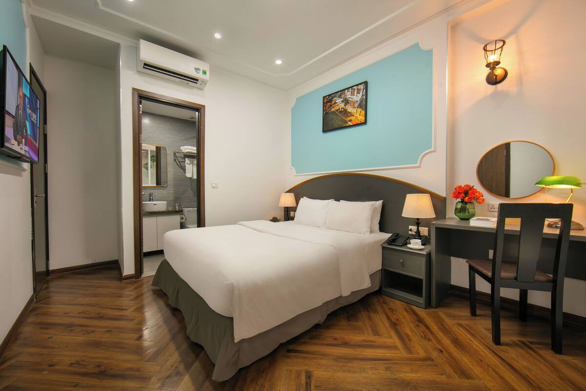 A hotel room with a bed, a desk with a chair, a desk lamp, a wall-mounted mirror, a picture on the wall, and an air conditioning unit.