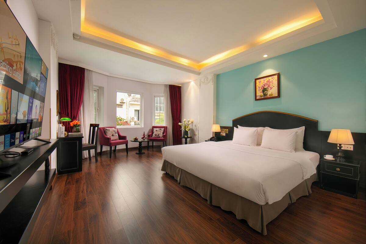 A hotel room with a large bed, a desk, a chair, a window with curtains, and a painting on the wall.