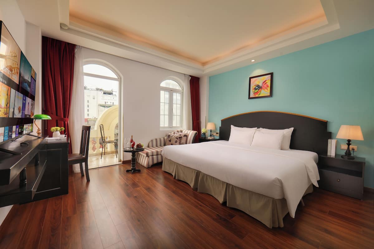 A hotel room with a large bed, a desk with a computer, a chair, a sofa, and a window with curtains.
