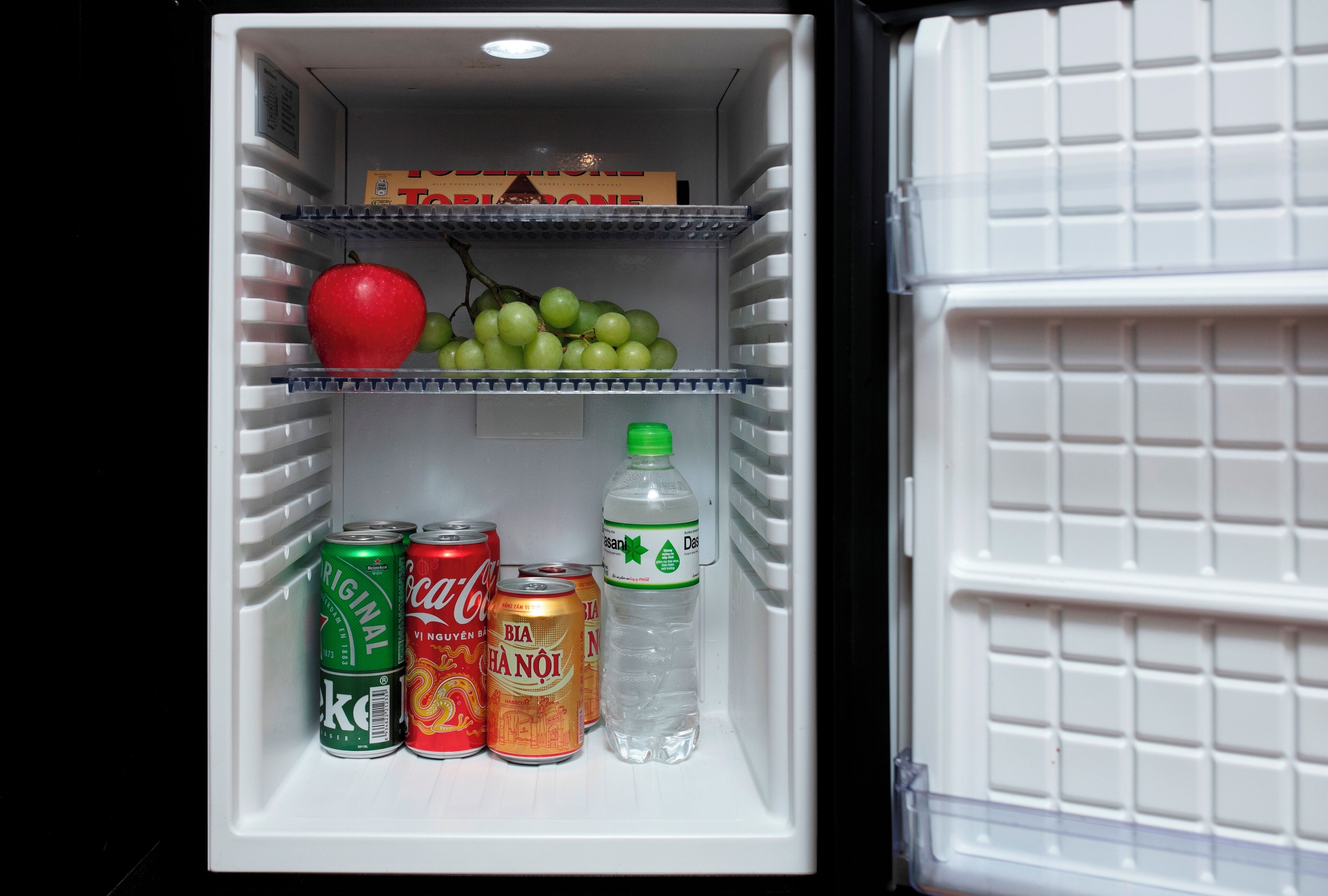 Un refrigerador con repisas que contienen una manzana roja, uvas verdes y varias latas de bebidas.