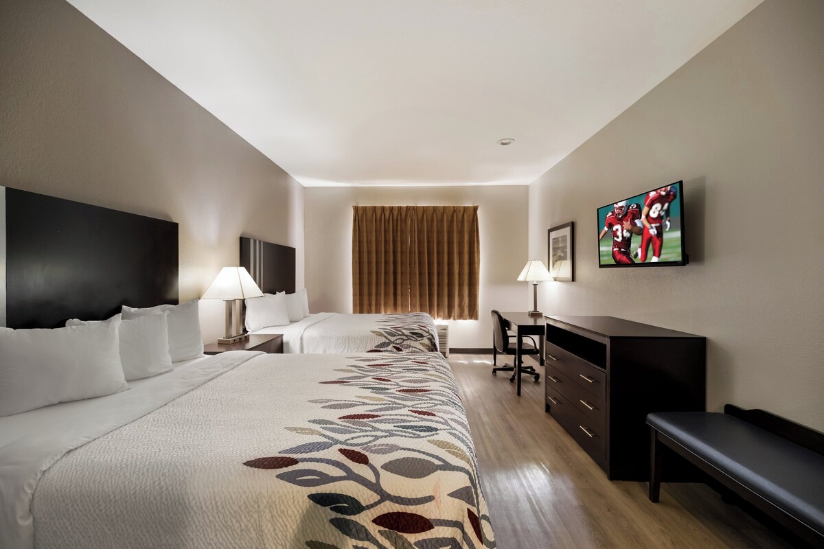 A hotel room with two beds, a desk, a chair, a TV, and a window with curtains.
