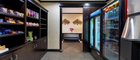A corridor with shelves stocked with snacks and beverages, a seating area with a vase of flowers, and a refrigerator with drinks.