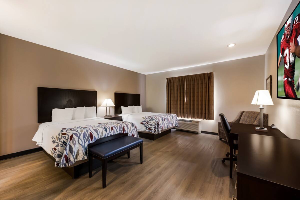 A hotel room with two beds, a desk, a chair, a bench, a TV, and a window with curtains.