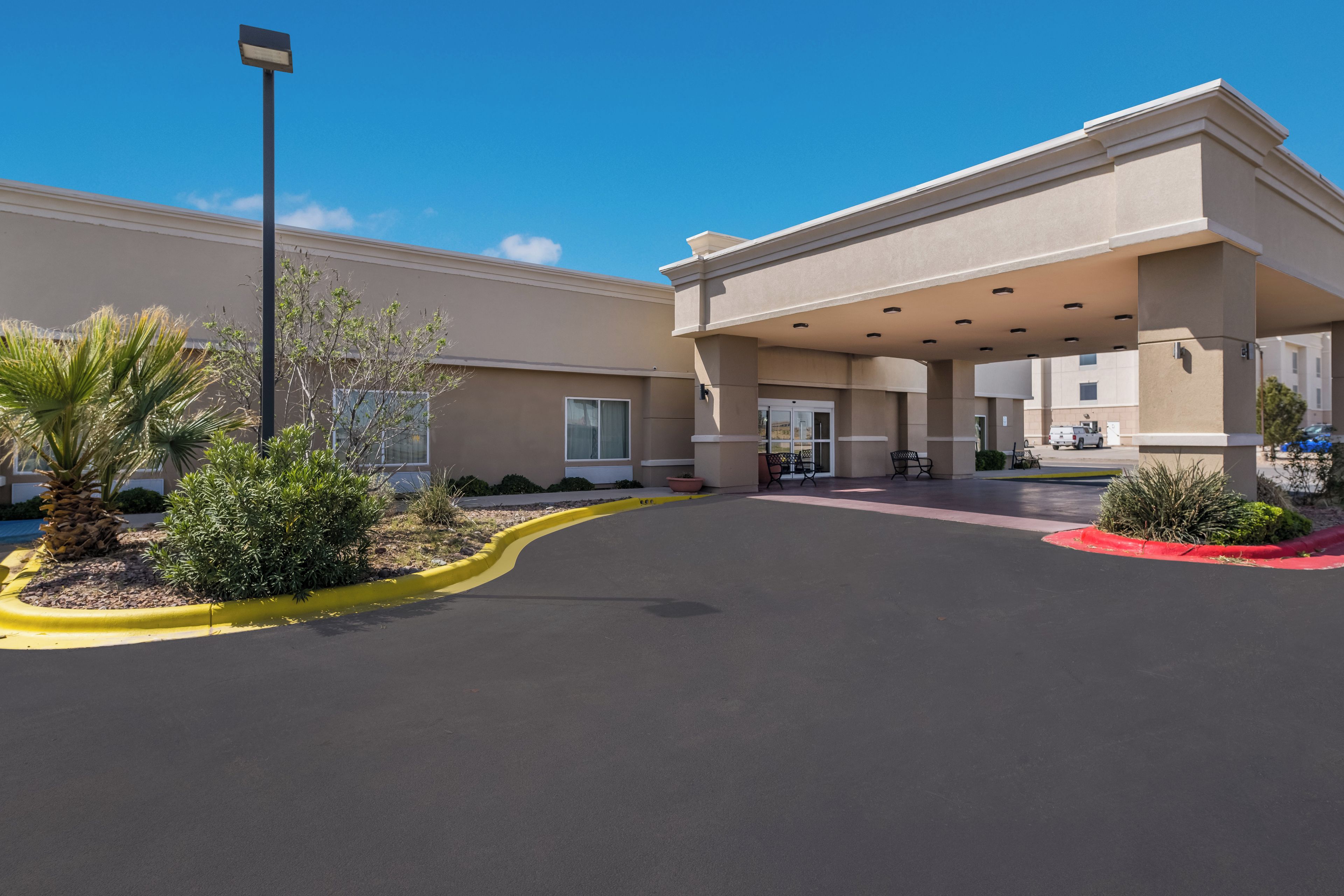 A hotel entrance with a covered waiting area, a parking lot, and landscaping.