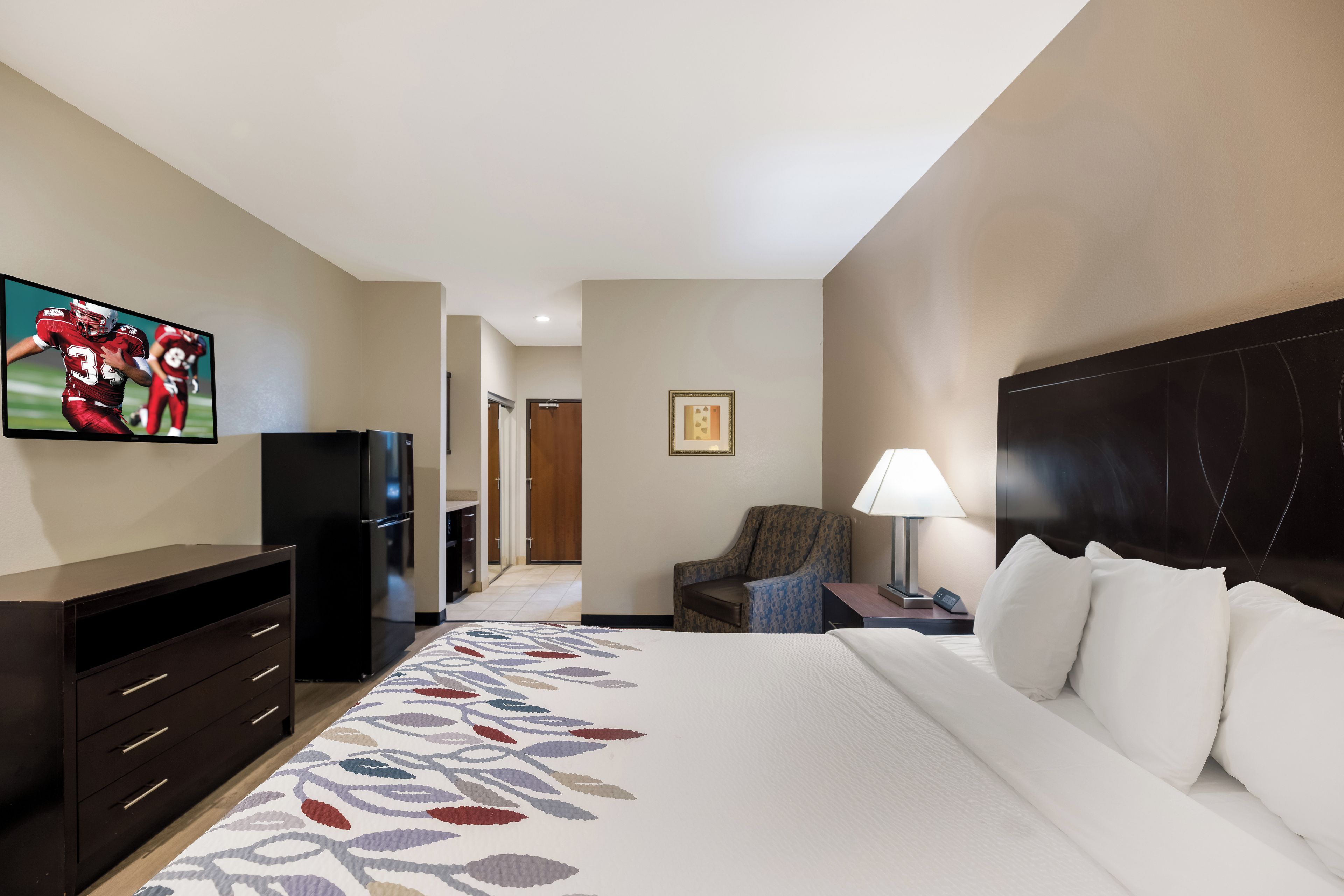 A hotel room with a bed, a TV, a desk with a lamp, a chair, and a refrigerator.