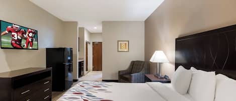 A hotel room with a bed, a TV, a desk with a lamp, a chair, and a refrigerator.