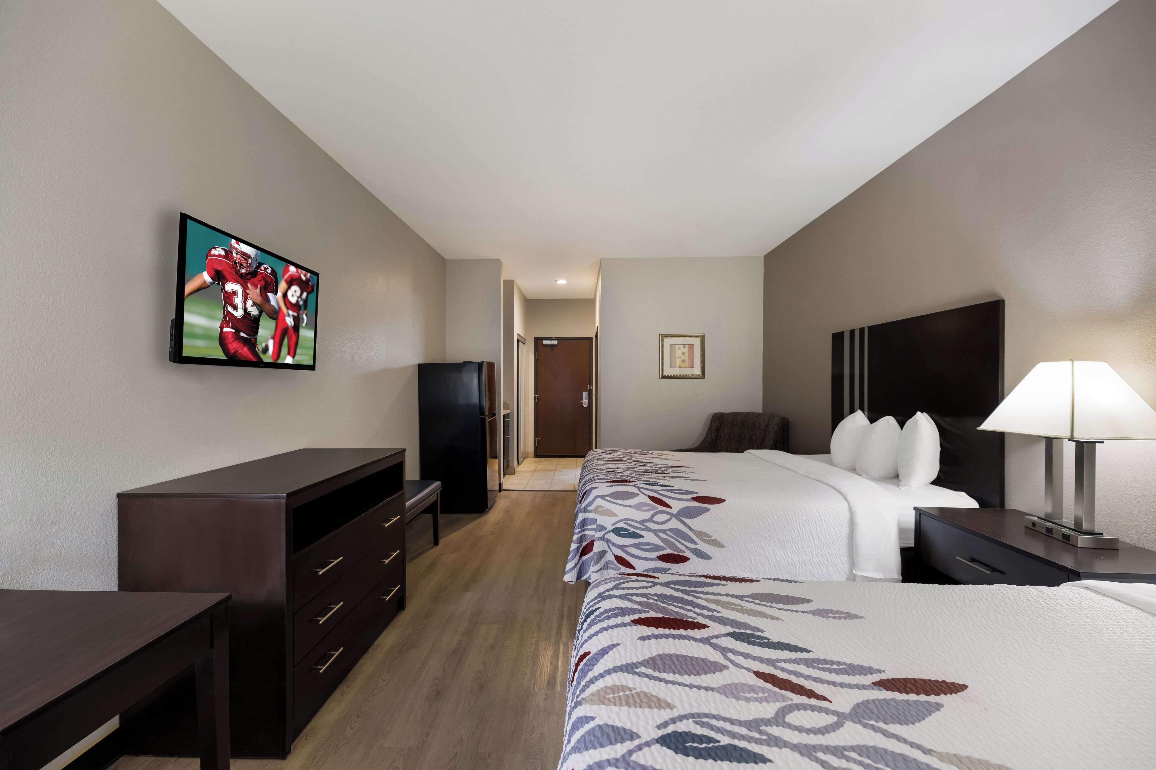 Una habitación de hotel con televisor de pantalla plana montado en la pared, cama con cubrecama estampado, mesita de noche con lámpara y cómoda con cajones.