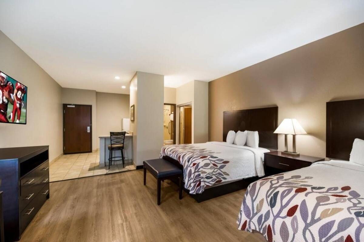 A hotel room with two beds, a desk, a chair, a TV, and a bathroom visible in the background.