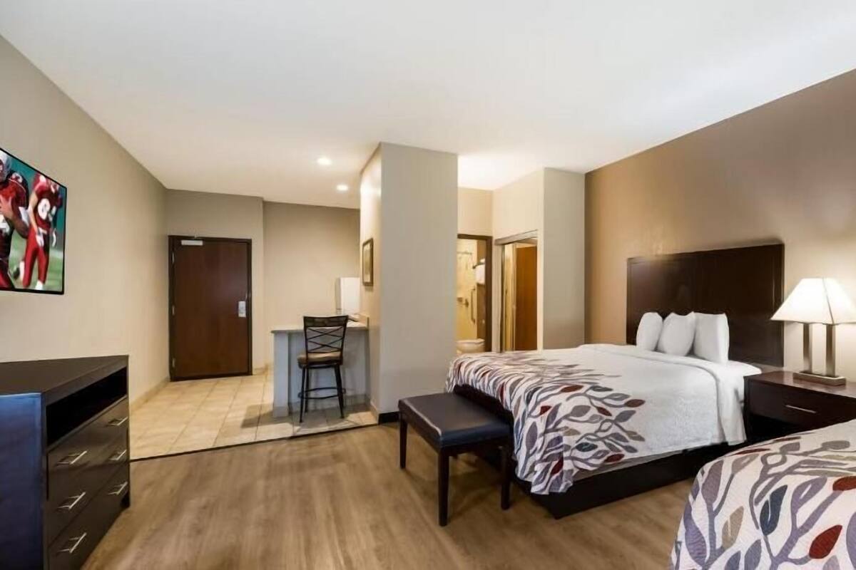 A hotel room with two beds, a desk, a chair, a TV, and a bathroom visible through an open door.