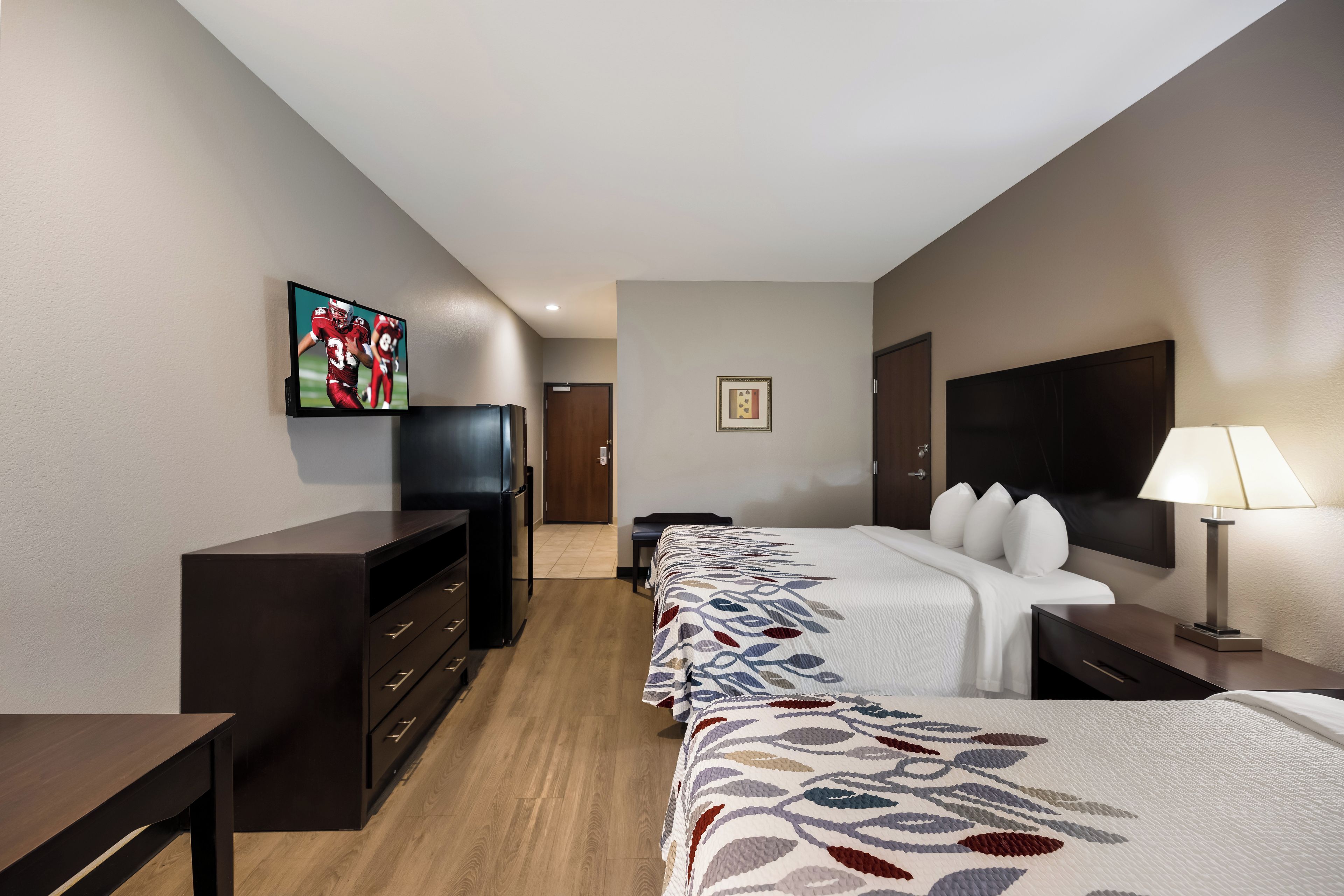 A hotel room with a bed, a desk, a television, a refrigerator, and a lamp.