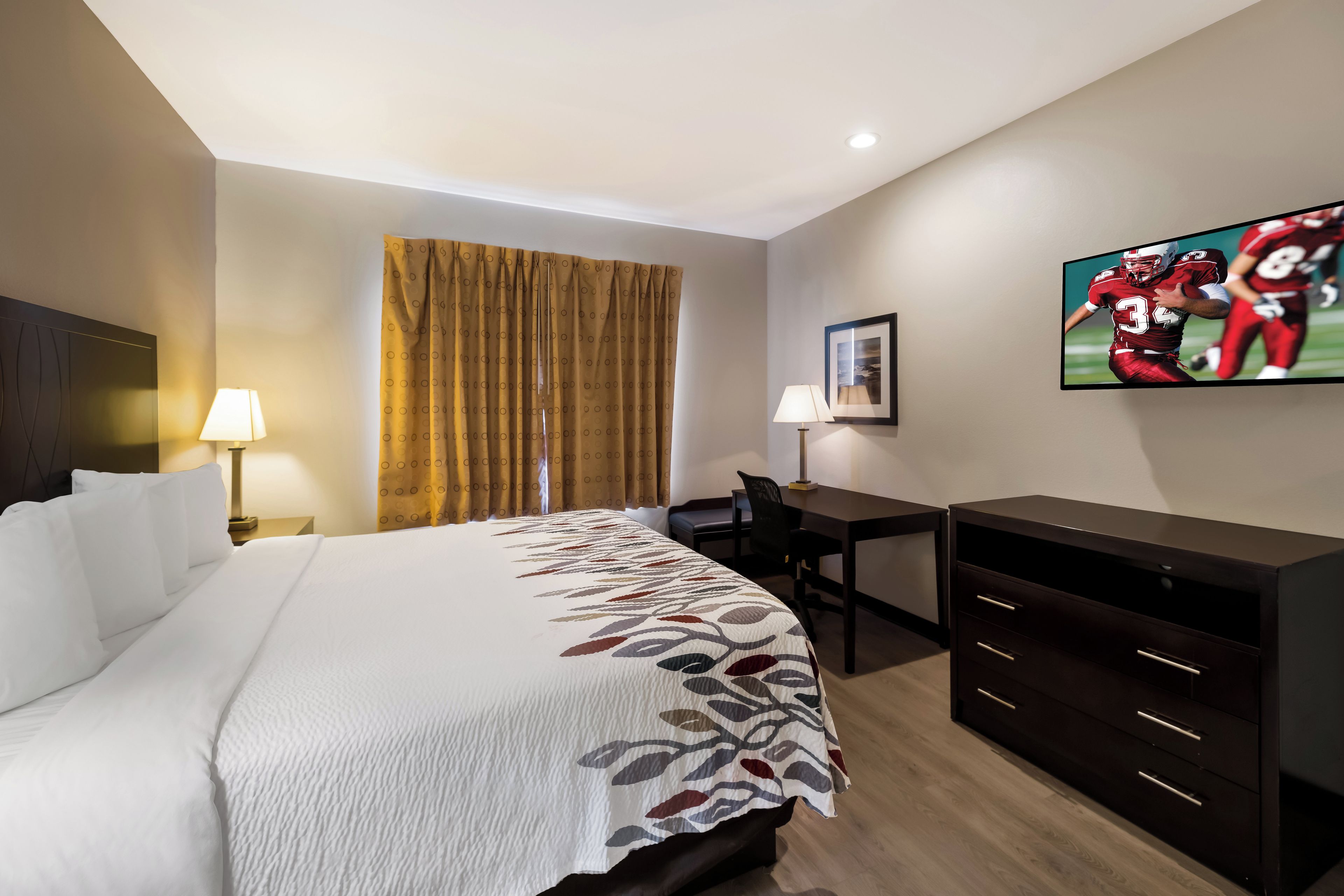 A hotel room with a bed, a desk, a chair, a TV, and a window with curtains.