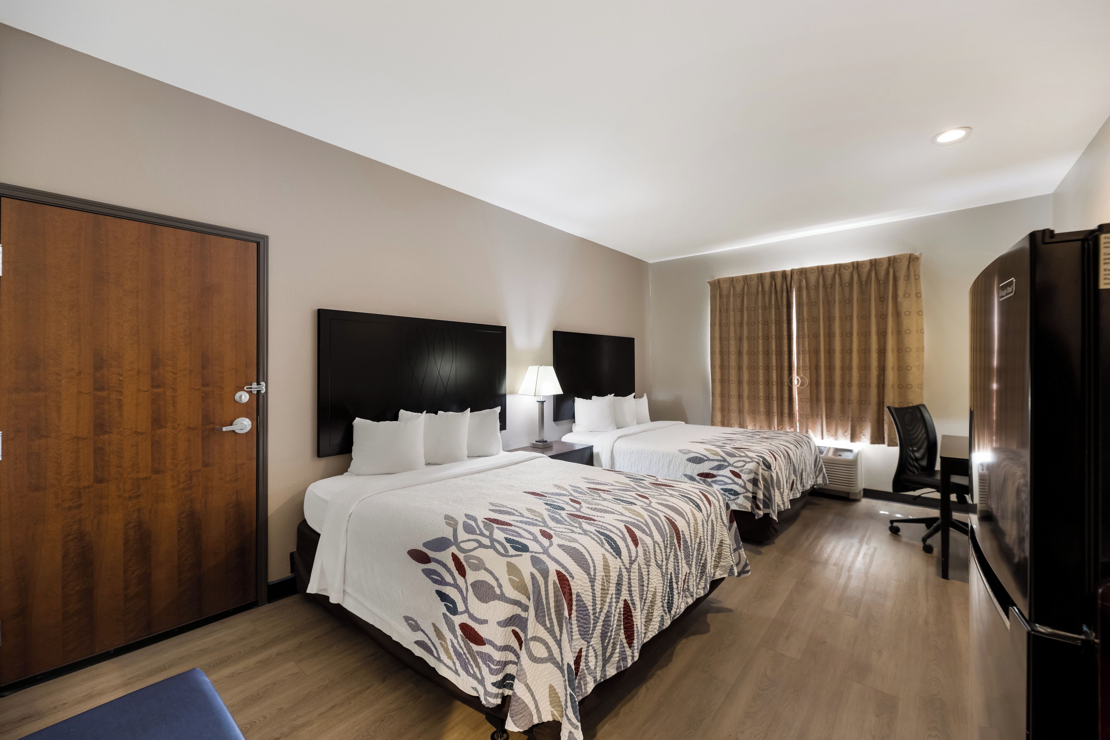 A hotel room with two beds, a wardrobe, a desk, a chair, a television, and a window with curtains.