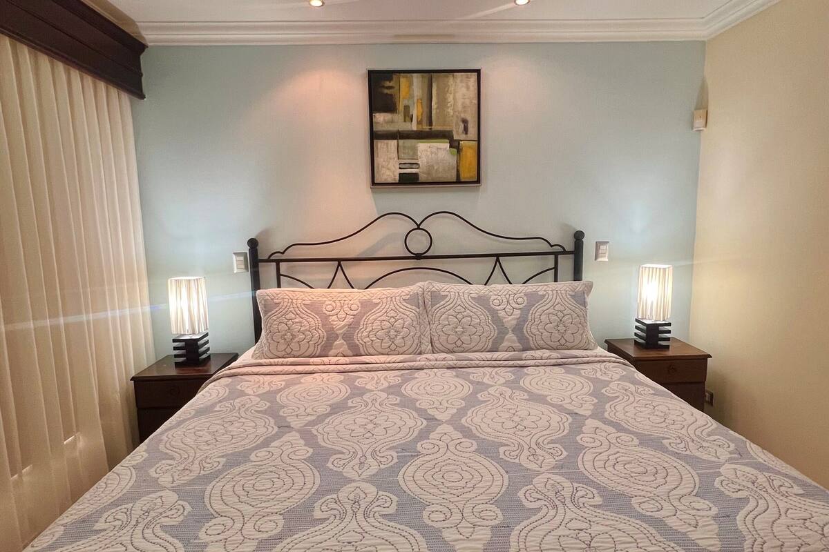 A bedroom with a bed, two bedside tables, a ceiling light, a wall-mounted painting, and a window with blinds.