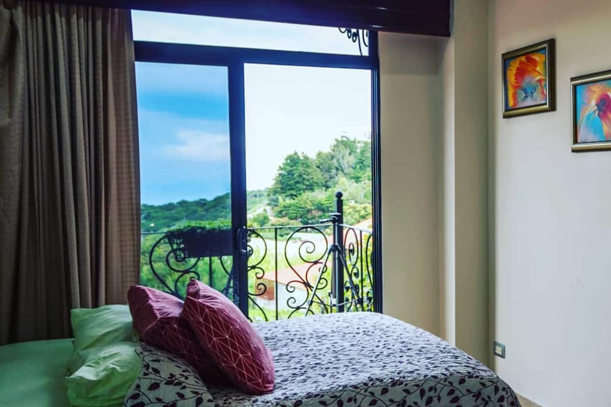 A bedroom with a large window, a bed with patterned bedding, and two framed artworks on the wall.