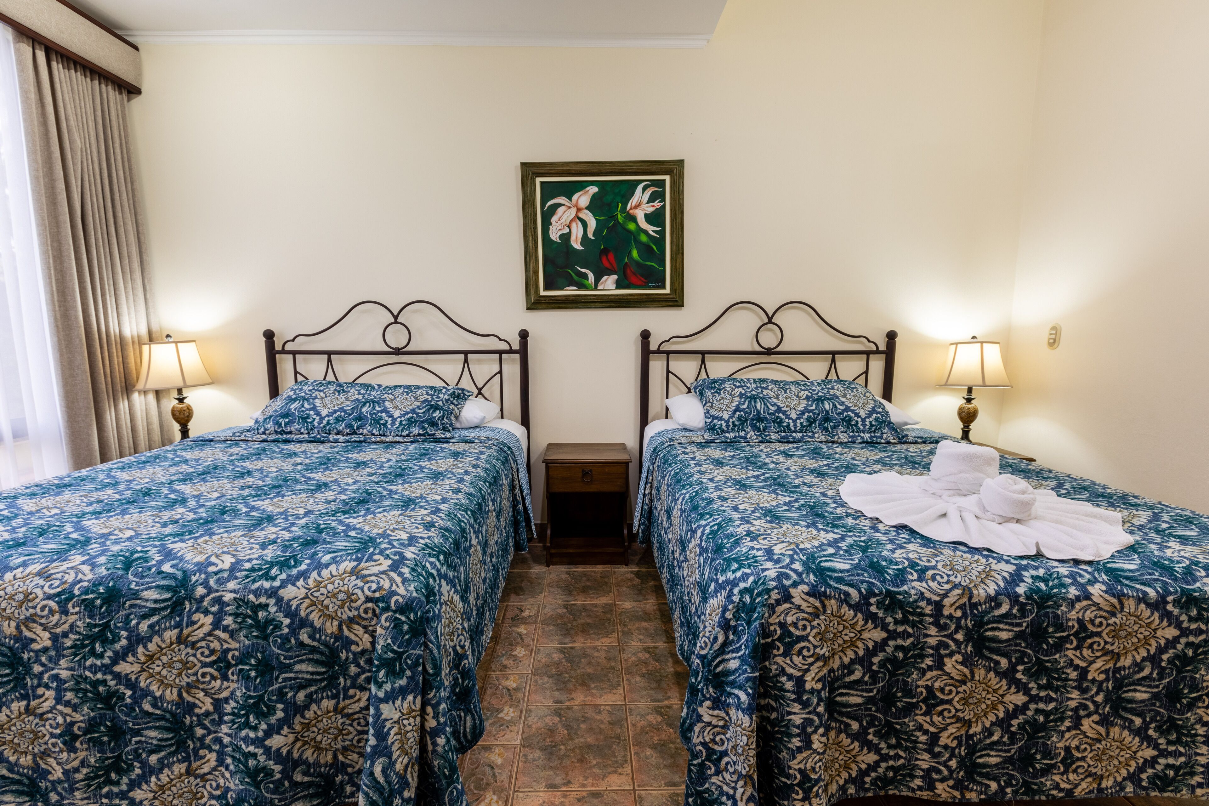 Quarto de hotel com duas camas, cada uma com colcha estampada em florais, uma mesa de cabeceira entre elas e um quadro na parede.