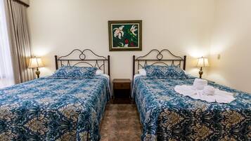 Quarto de hotel com duas camas, cada uma com colcha estampada em florais, uma mesa de cabeceira entre elas e um quadro na parede.
