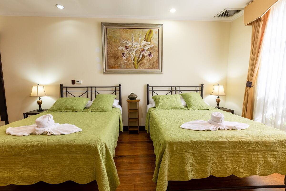 A hotel room with two beds, each with a green bedspread and pillows, a small table with a lamp, and a painting of flowers on the wall.
