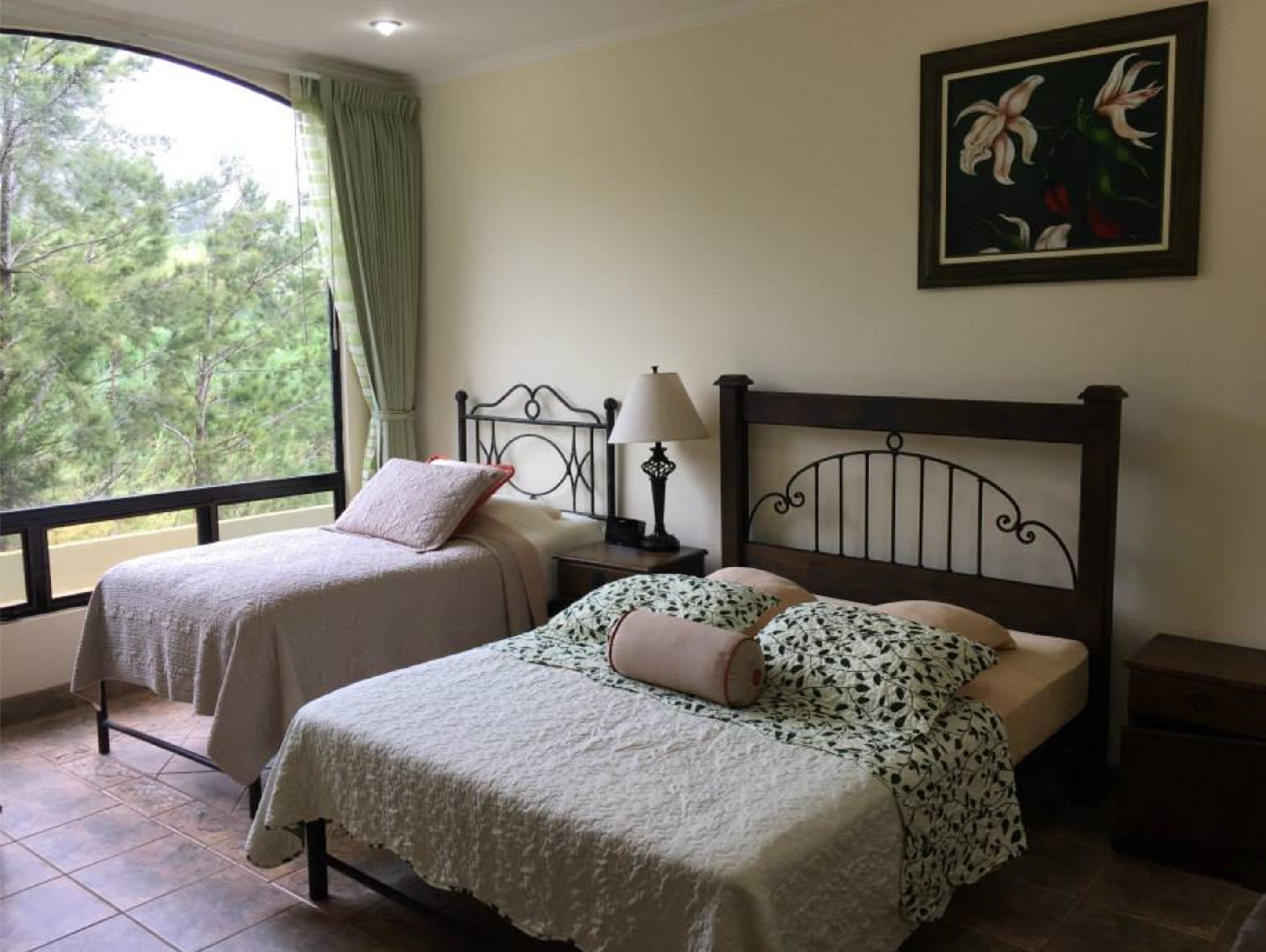 Una habitación con dos camas, una ventana que da a árboles, un cuadro en la pared y una lámpara en la mesita de noche.
