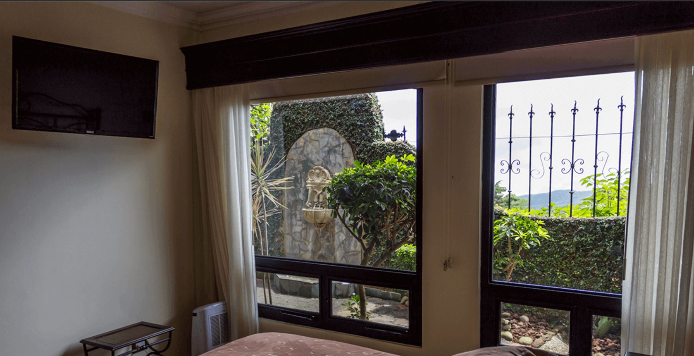 Een slaapkamer met een bed, een klein tafeltje en een raam met uitzicht op een tuin en een standbeeld.