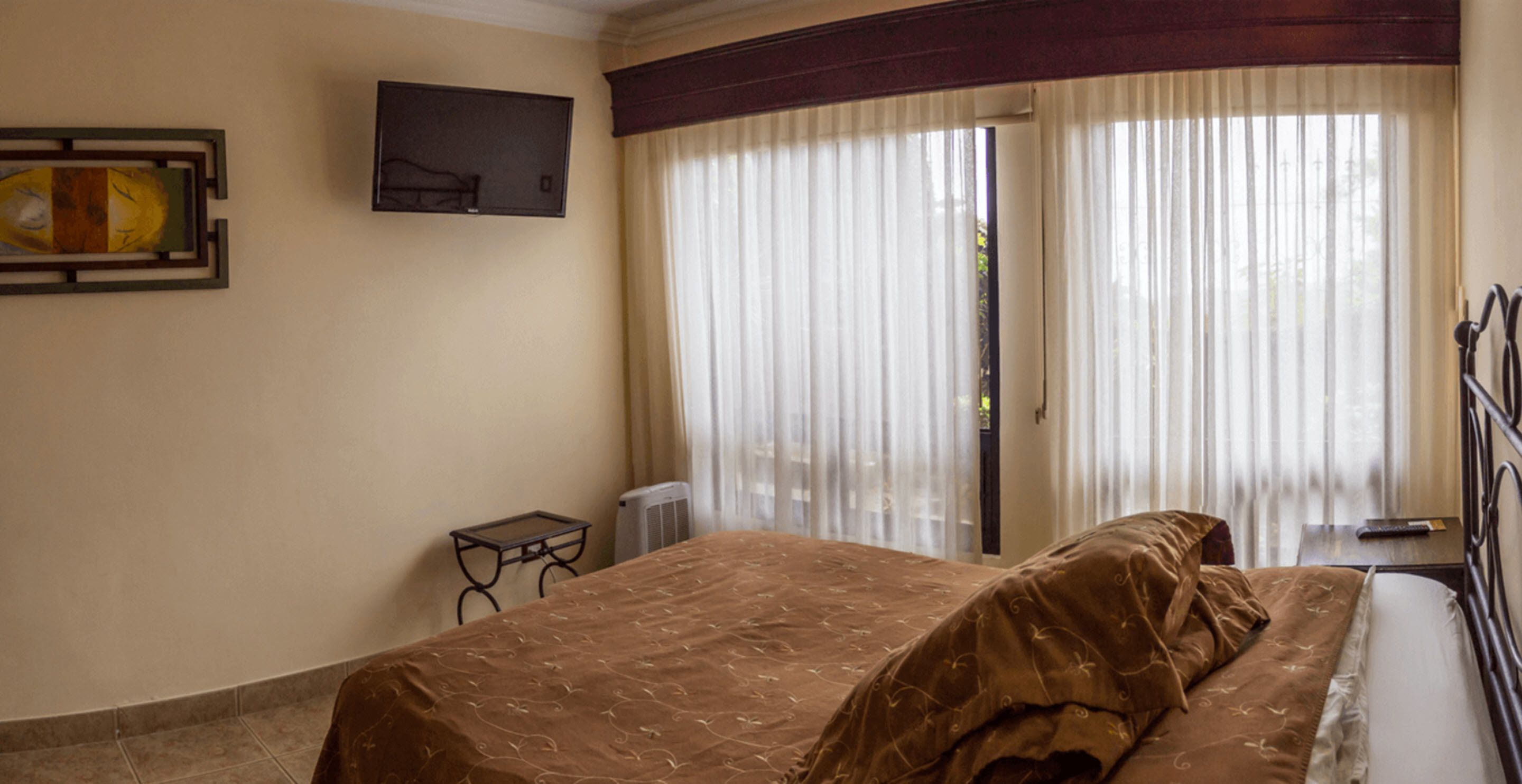 Una habitación de hotel con cama, televisión en la pared y ventana con cortinas.