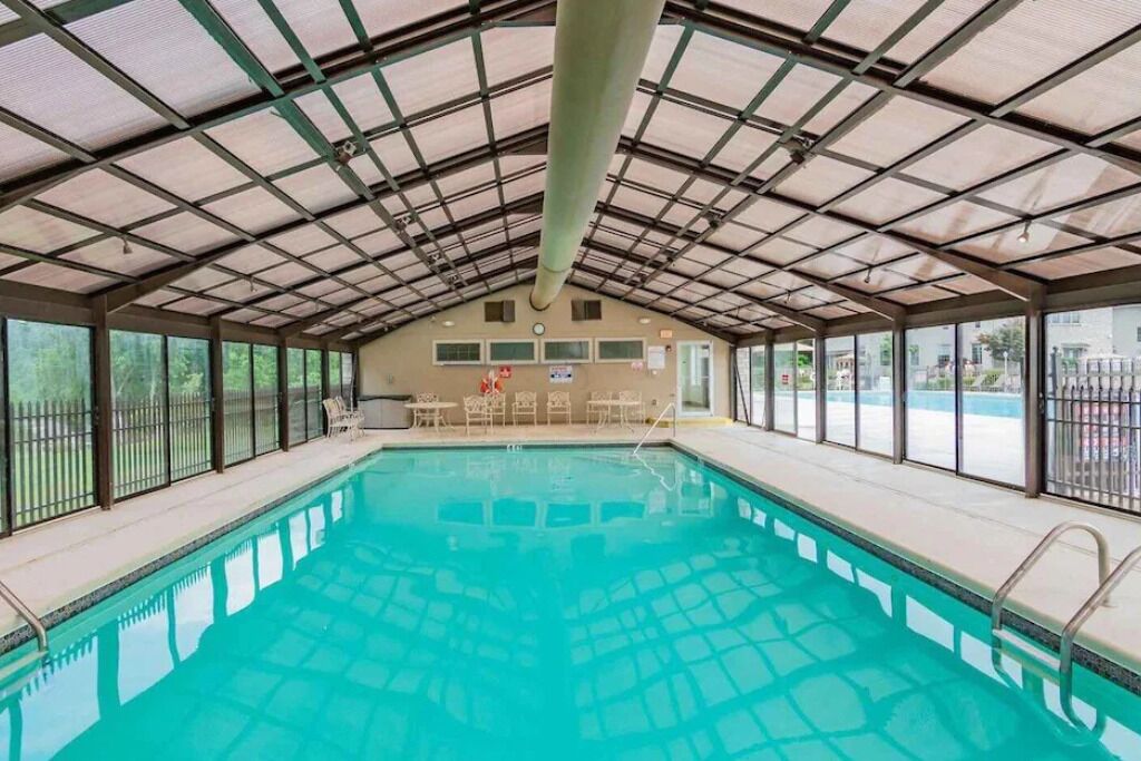An indoor swimming pool with a metal roof and glass walls, surrounded by lounge chairs and tables.