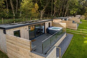 Una cabaña de madera moderna con balcón de vidrio, un jacuzzi y vistas del bosque circundante.