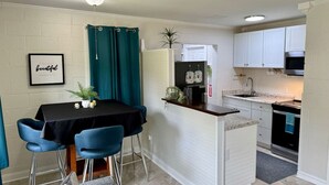 Een kleine keuken met witte kastjes, een zwart aanrecht en een eilandje. Er is een eethoek met een zwarte tafel en blauwe stoelen.