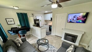 Een woonkamer met grijze banken, een salontafel, een televisie, een open haard en een keuken op de achtergrond.