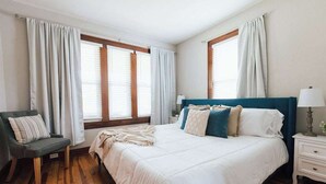 Un dormitorio con una cama grande, una silla, una mesita de noche y dos ventanas con cortinas.
